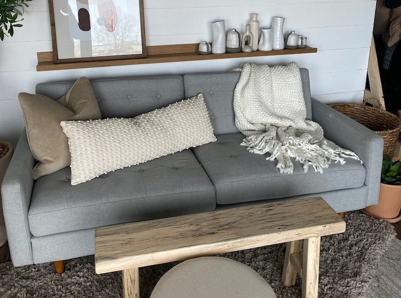 May include: A gray sofa with a white throw blanket and pillows. The sofa is in front of a white wall with a wooden shelf above it. There are white ceramic vases on the shelf. A wooden bench is in front of the sofa.