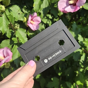 May include: A black plastic cassette tape keychain with a Spotify code printed on it. The keychain is being held in a hand against a backdrop of green leaves and pink flowers.