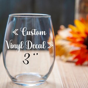 May include: A clear glass wine glass with a white vinyl decal that says "Custom Vinyl Decal" with arrows pointing in and out and "3" with quotation marks.