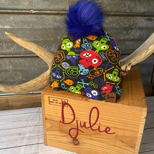 Peut inclure: Un bonnet noir avec un pompon bleu et un motif coloré de crânes et d'os croisés. Le bonnet porte l'inscription "Bad". Le bonnet est posé sur une boîte en bois sur laquelle est inscrit le mot "Julie".
