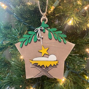 May include: A wooden star-shaped Christmas ornament with a green leafy design at the top. The ornament depicts a yellow manger with a white baby inside and a yellow star above.