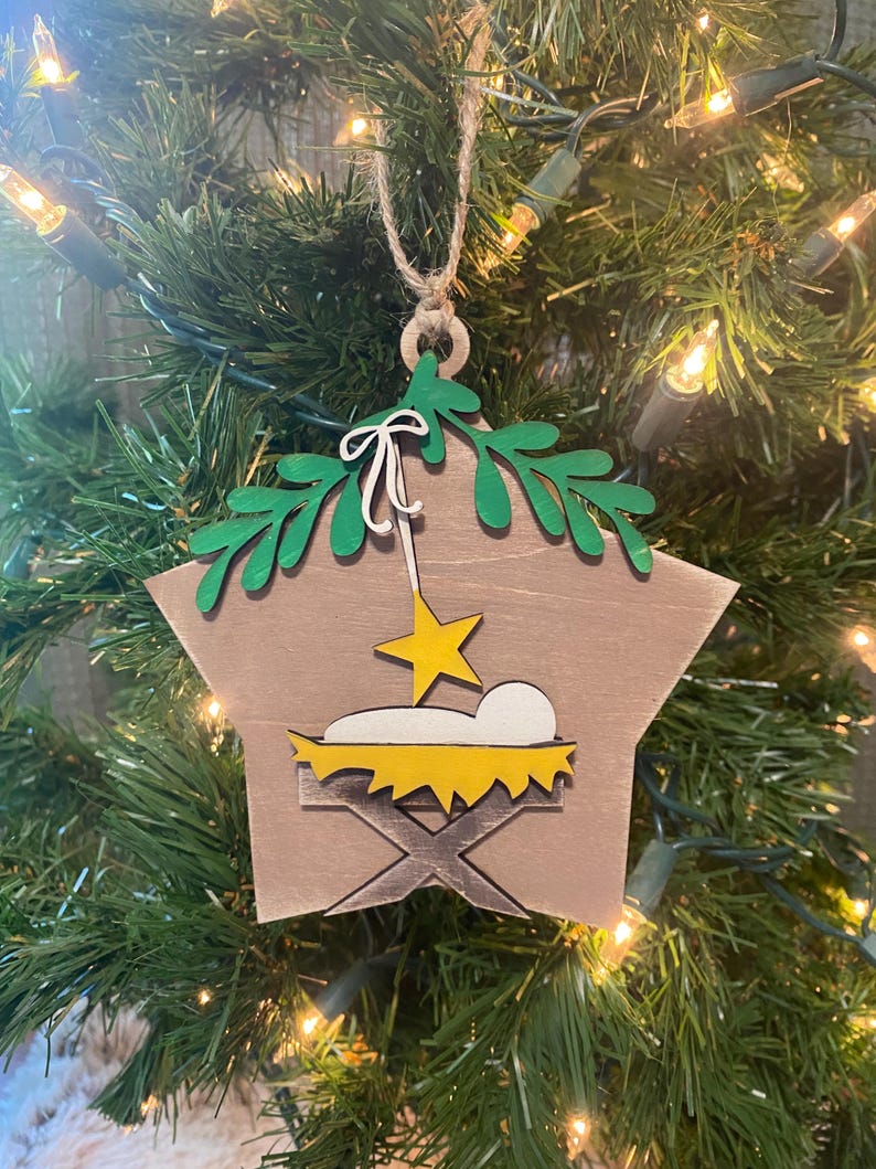 May include: A wooden star-shaped Christmas ornament with a green leafy design at the top. The ornament depicts a yellow manger with a white baby inside and a yellow star above.