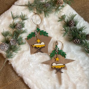 May include: Two wooden star-shaped Christmas ornaments with a nativity scene design. Each ornament features a green wreath, a yellow star, and a brown manger with a white baby Jesus inside.