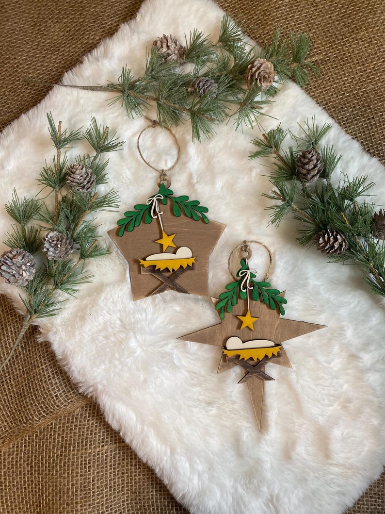 May include: Two wooden star-shaped Christmas ornaments with a nativity scene design. Each ornament features a green wreath, a yellow star, and a brown manger with a white baby Jesus inside.