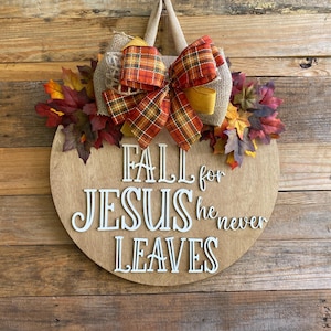 May include: A round wooden sign with the text "Fall for Jesus he never leaves" in white lettering. The sign is decorated with a burlap bow, a plaid ribbon, and fall leaves.