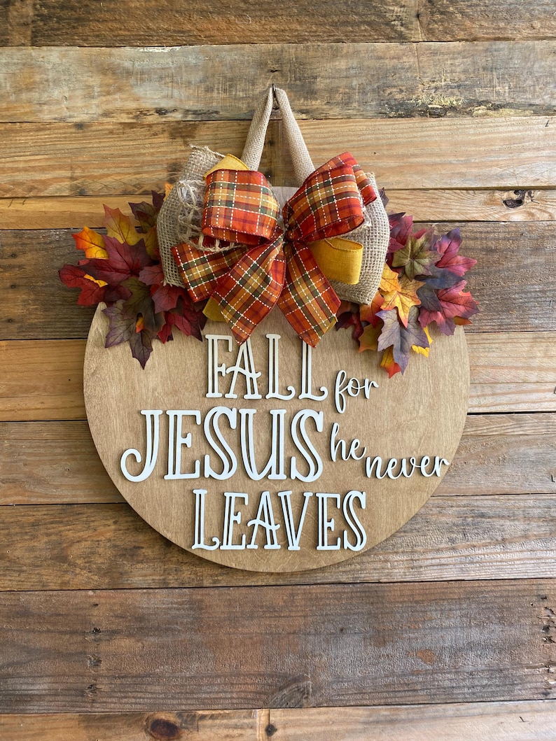 May include: A round wooden sign with the text "Fall for Jesus he never leaves" in white lettering. The sign is decorated with a burlap bow, a plaid ribbon, and fall leaves.
