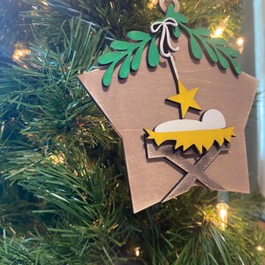 May include: A wooden star-shaped Christmas ornament with a brown background. The ornament features a green palm tree, a yellow star, and a yellow manger with a white baby Jesus inside.