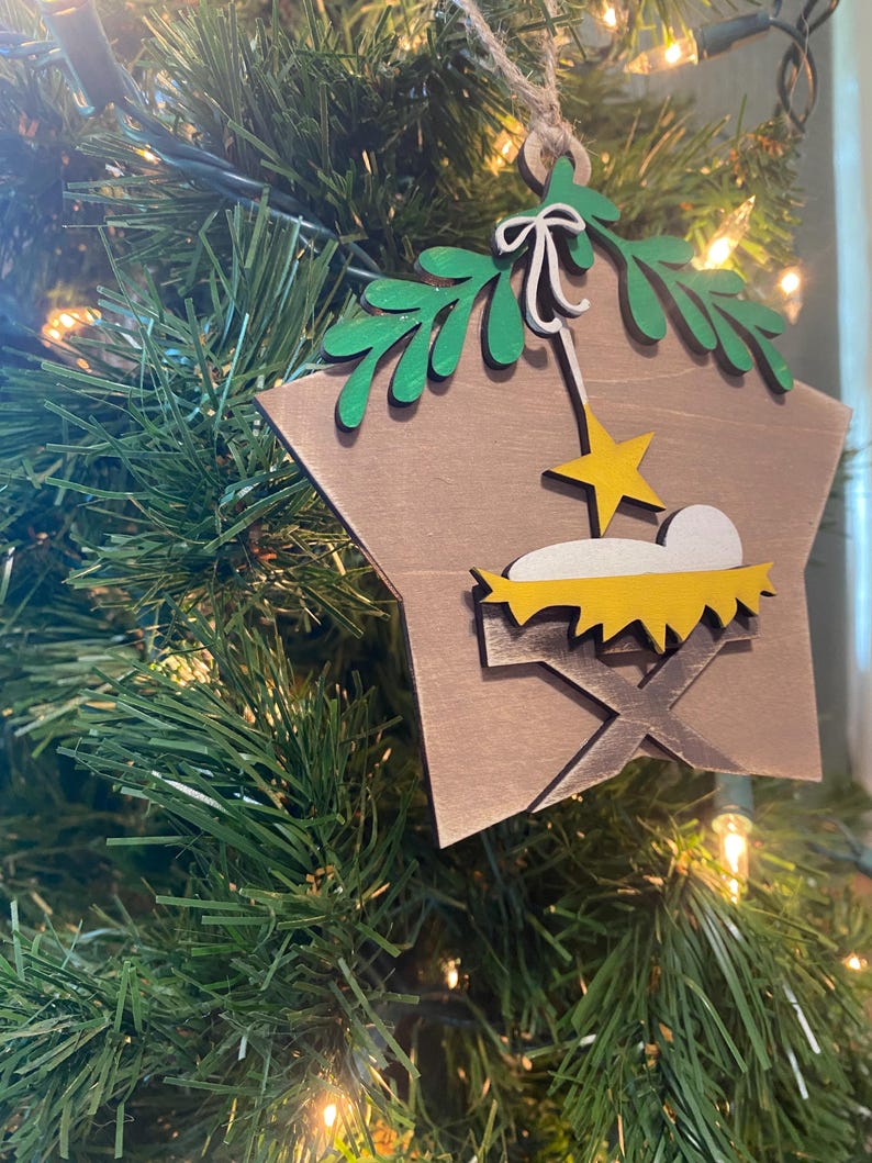 May include: A wooden star-shaped Christmas ornament with a brown background. The ornament features a green palm tree, a yellow star, and a yellow manger with a white baby Jesus inside.