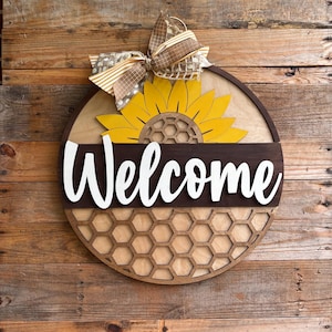 May include: A round wooden welcome sign with a sunflower and honeycomb design. The sign features the word "Welcome" in white script, a yellow sunflower, and a decorative bow. The sign is made of wood and has a rustic look.