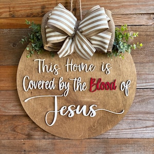 May include: A round wooden door sign featuring the white text This Home is Covered by the Blood of Jesus with the word Blood in red. The sign is decorated with a beige and white striped burlap bow and green artificial foliage at the top.