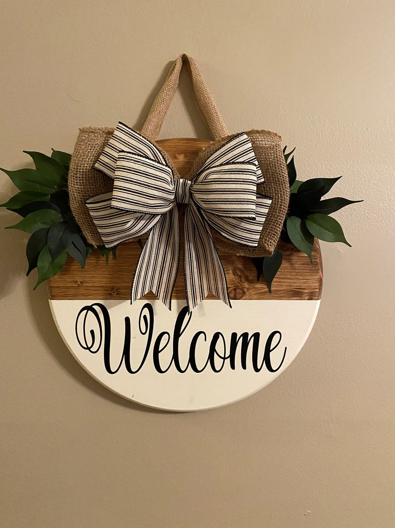 May include: A round wooden door hanger with a white and brown painted design. The hanger has a burlap bow with black and white stripes and green leaves. The words "Welcome" are written in black on the white portion of the hanger.