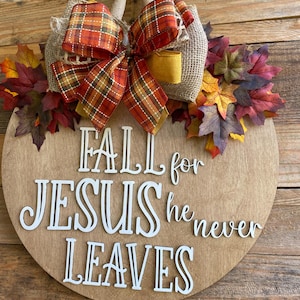 May include: A round wooden sign with the text "FALL for JESUS he never LEAVES" in white letters. The sign is decorated with a burlap bow, a plaid ribbon, and fall leaves.