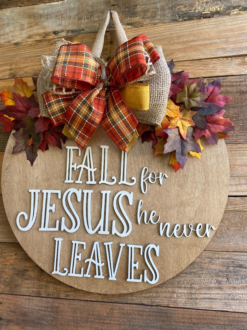 May include: A round wooden sign with the text "FALL for JESUS he never LEAVES" in white letters. The sign is decorated with a burlap bow, a plaid ribbon, and fall leaves.