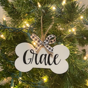 May include: White bone-shaped Christmas ornament with the name "Grace" written in black calligraphy. The ornament is decorated with a black and white checkered ribbon.