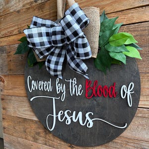 May include: A round, dark wooden sign with the words "Covered by the Blood of Jesus" in white and red lettering. The sign is decorated with a black and white checkered bow, burlap, and green leaves. A jute loop is attached for hanging.