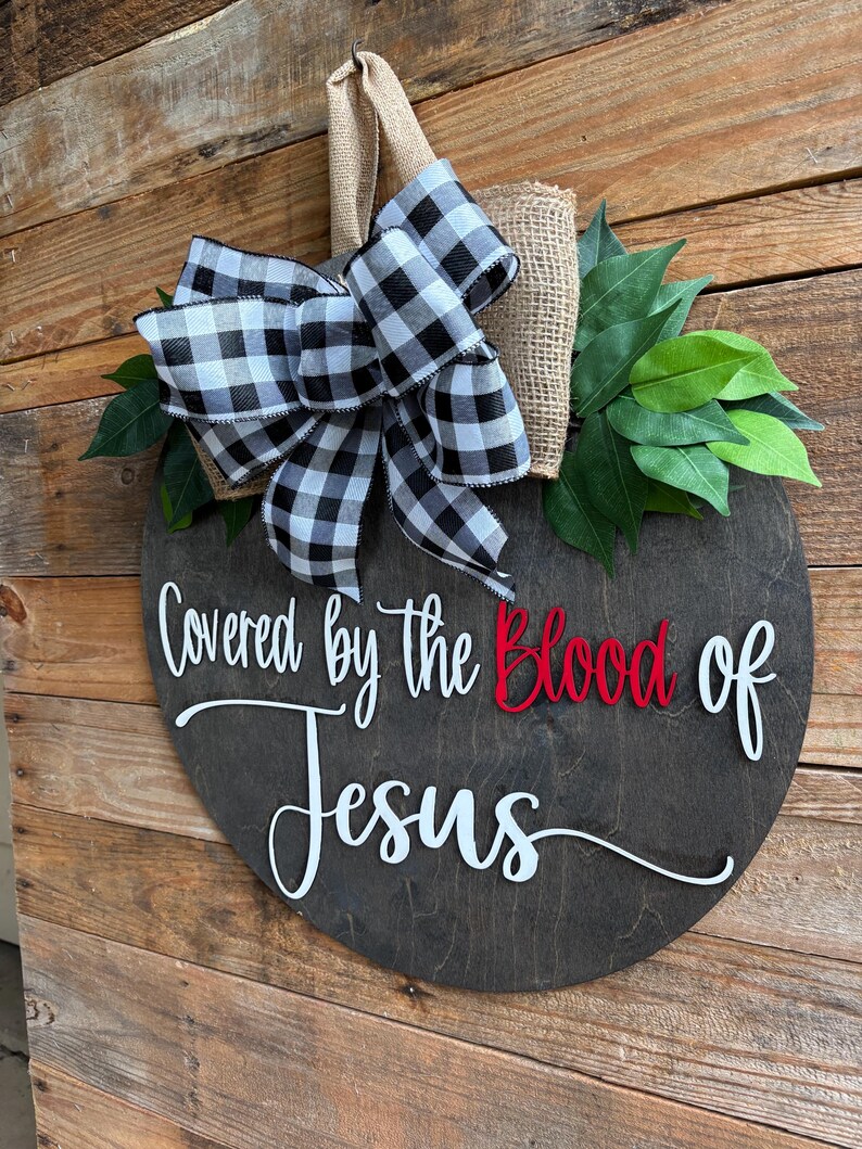 May include: A round, dark wooden sign with the words "Covered by the Blood of Jesus" in white and red lettering. The sign is decorated with a black and white checkered bow, burlap, and green leaves. A jute loop is attached for hanging.