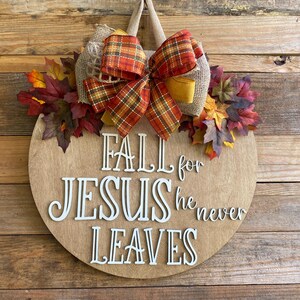 May include: A round wooden door hanger with a burlap bow and fall leaves. The sign reads "Fall for Jesus he never leaves".