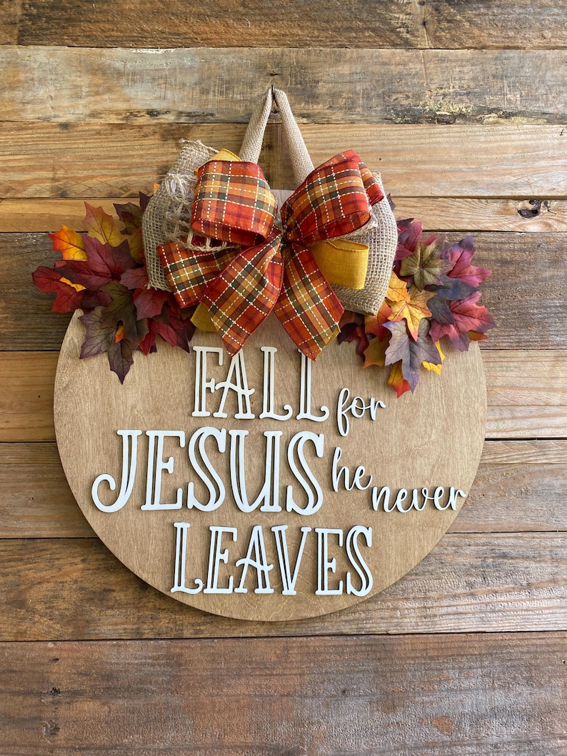 May include: A round wooden door hanger with a burlap bow and fall leaves. The sign reads "Fall for Jesus he never leaves".