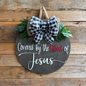 May include: A round wooden sign with the text "Covered by the Blood of Jesus" in white and red lettering. The sign is decorated with a black and white checkered bow, burlap, and green leaves. It hangs from a jute loop.