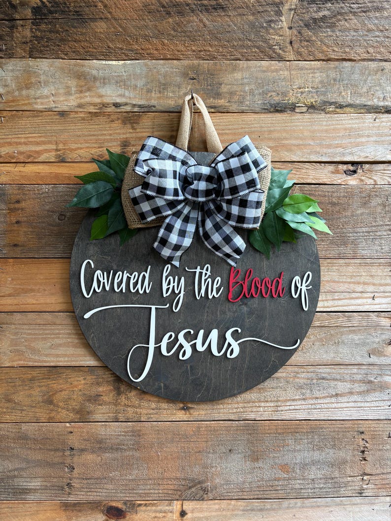 May include: A round wooden sign with the text "Covered by the Blood of Jesus" in white and red lettering. The sign is decorated with a black and white checkered bow, burlap, and green leaves. It hangs from a jute loop.