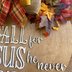 May include: A wooden round sign with the words "Fall for Jesus he never leaves" in white letters. The sign is decorated with a burlap bow, a yellow ribbon, and fall leaves.
