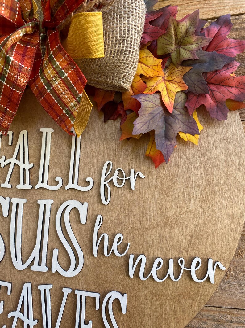 May include: A wooden round sign with the words "Fall for Jesus he never leaves" in white letters. The sign is decorated with a burlap bow, a yellow ribbon, and fall leaves.