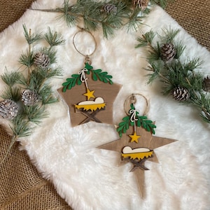 May include: Two wooden star-shaped Christmas ornaments with a nativity scene design. The ornaments are painted brown with green leaves, a yellow star, and a yellow manger with a white baby Jesus inside.