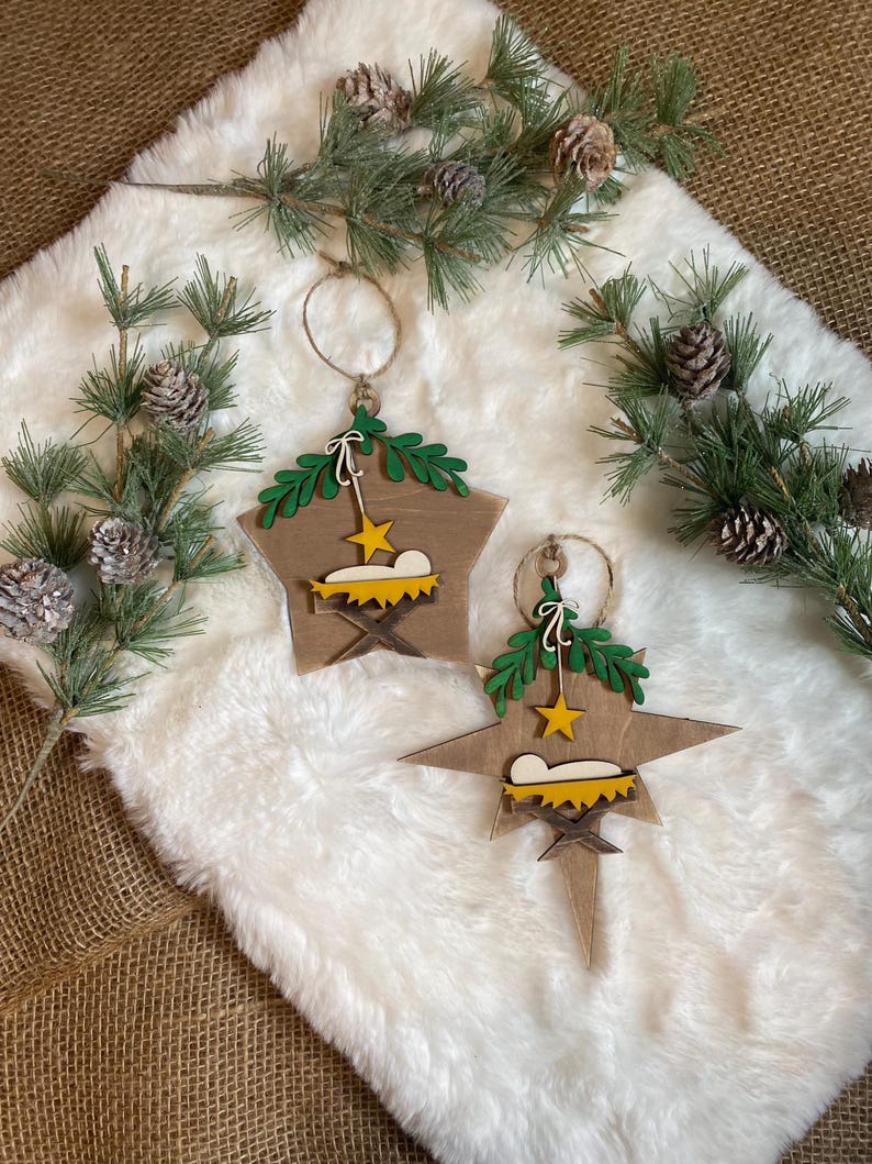 May include: Two wooden star-shaped Christmas ornaments with a nativity scene design. The ornaments are painted brown with green leaves, a yellow star, and a yellow manger with a white baby Jesus inside.