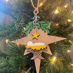May include: A wooden star-shaped Christmas ornament with a green holly wreath, a yellow star, and a yellow manger with a white baby Jesus inside.