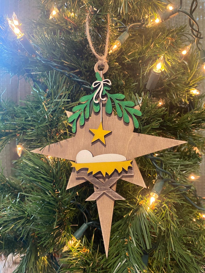 May include: A wooden star-shaped Christmas ornament with a green holly wreath, a yellow star, and a yellow manger with a white baby Jesus inside.