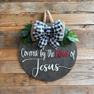 May include: Round wooden sign with the text "Covered by the Blood of Jesus" in white and red lettering. The sign is decorated with a black and white checkered bow, burlap, and green leaves. It hangs from a jute rope.
