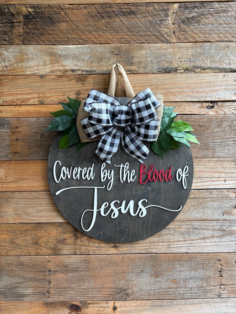May include: Round wooden sign with the text "Covered by the Blood of Jesus" in white and red lettering. The sign is decorated with a black and white checkered bow, burlap, and green leaves. It hangs from a jute rope.