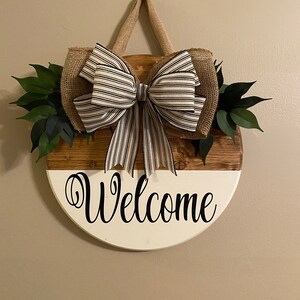 May include: A round wooden welcome sign with a white and brown wood grain finish. The sign has a black and white striped bow and green leaves attached to it. The sign says "Welcome" in black cursive lettering.