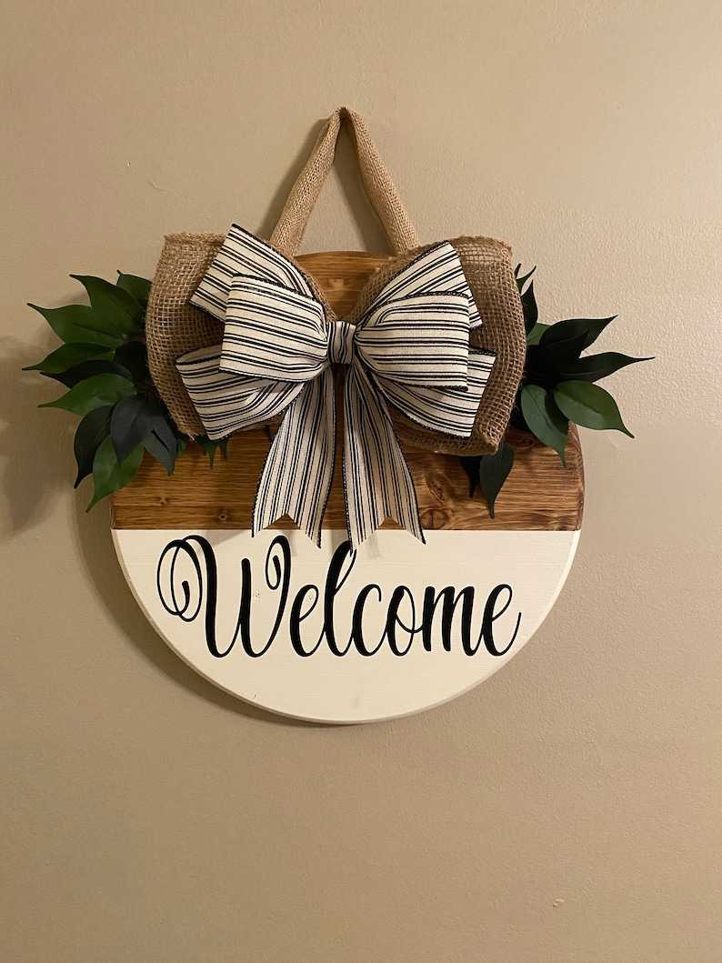 May include: A round wooden welcome sign with a white and brown wood grain finish. The sign has a black and white striped bow and green leaves attached to it. The sign says "Welcome" in black cursive lettering.