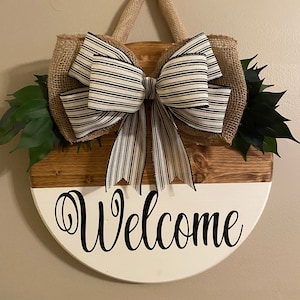May include: A round wooden welcome sign with a white and brown painted finish. The sign has a burlap bow with black and white striped ribbon and green leaves. The sign says "Welcome" in black cursive lettering.