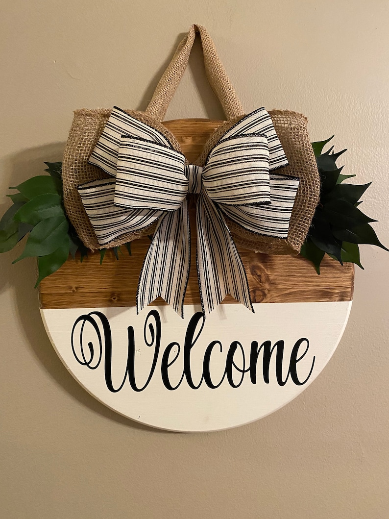 May include: A round wooden welcome sign with a white and brown painted finish. The sign has a burlap bow with black and white striped ribbon and green leaves. The sign says "Welcome" in black cursive lettering.