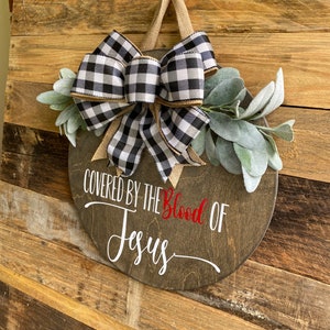 May include: A round wooden sign with the text "COVERED BY THE Blood OF Jesus" in white and red. It is decorated with a black and white buffalo check bow, burlap ribbon, and faux greenery. A jute loop is attached for hanging.