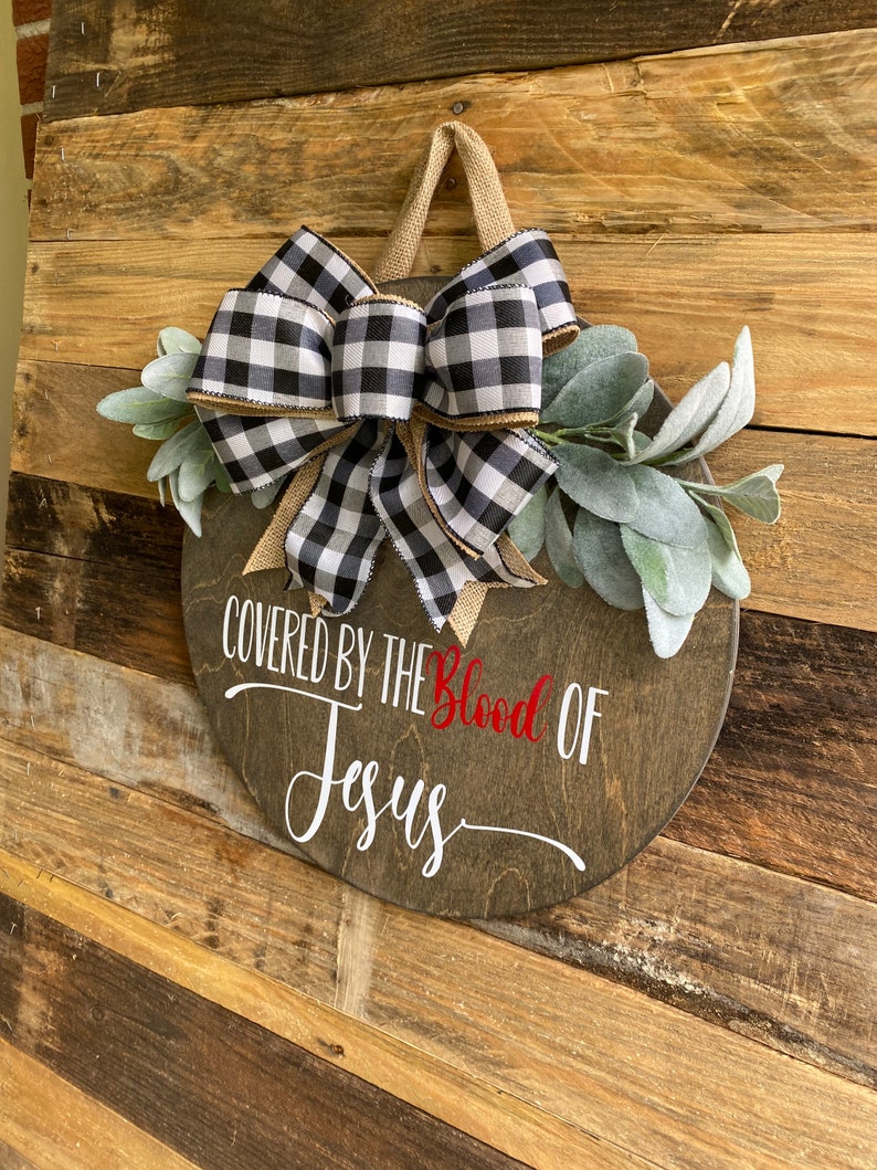 May include: A round wooden sign with the text "COVERED BY THE Blood OF Jesus" in white and red. It is decorated with a black and white buffalo check bow, burlap ribbon, and faux greenery. A jute loop is attached for hanging.