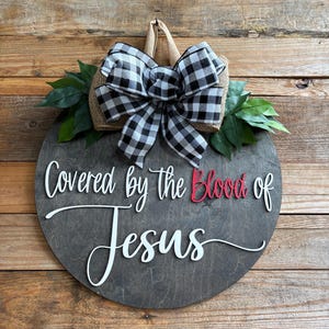 May include: A round wooden sign with the text "Covered by the Blood of Jesus" in white script and red lettering. The sign is decorated with a black and white checkered bow, burlap, and green leaves.