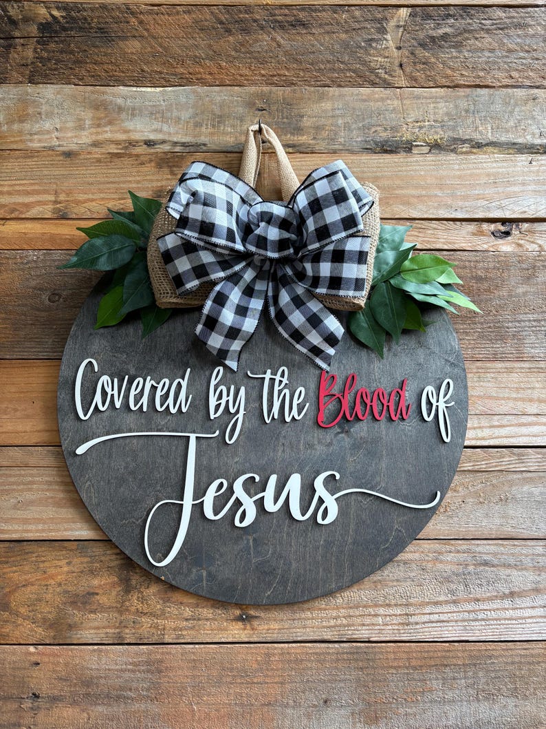 May include: A round wooden sign with the text "Covered by the Blood of Jesus" in white script and red lettering. The sign is decorated with a black and white checkered bow, burlap, and green leaves.