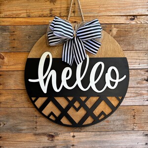 May include: Round wooden sign with the word "hello" in white script. The top half is natural wood, the bottom half is black with a geometric design. A black and white striped bow hangs from a jute string.