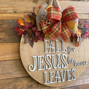 May include: A round wooden sign with the words "Fall for Jesus he never leaves" in white letters. The sign is decorated with a plaid bow, burlap, and fall leaves.