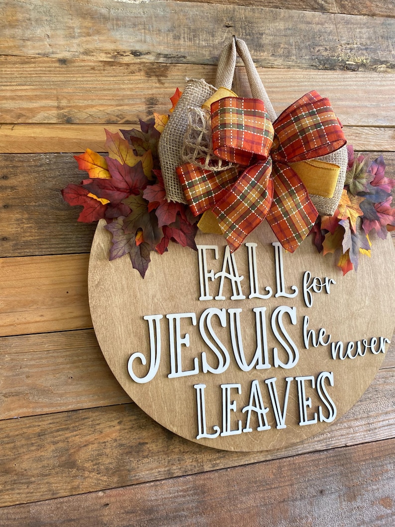 May include: A round wooden sign with the words "Fall for Jesus he never leaves" in white letters. The sign is decorated with a plaid bow, burlap, and fall leaves.