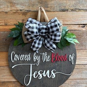 May include: A round wooden sign with the text "Covered by the Blood of Jesus" in white and red. It features a burlap and black and white checkered bow, green leaves, and a jute hanger. The sign is displayed against a wooden background.