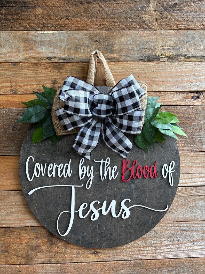 May include: A round wooden sign with the text "Covered by the Blood of Jesus" in white and red. It features a burlap and black and white checkered bow, green leaves, and a jute hanger. The sign is displayed against a wooden background.