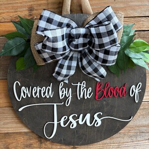 May include: A round, dark stained wooden sign with the text "Covered by the Blood of Jesus" in white and red. It features a large black and white checkered bow, burlap backing, and green leaves.