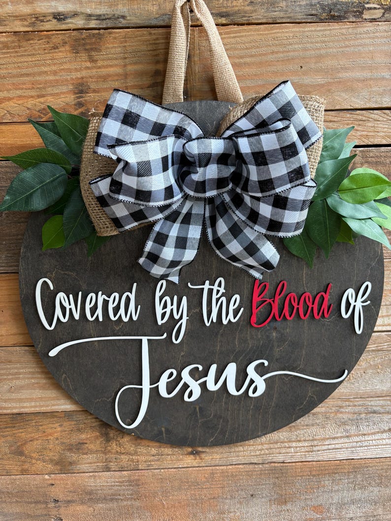 May include: A round, dark stained wooden sign with the text "Covered by the Blood of Jesus" in white and red. It features a large black and white checkered bow, burlap backing, and green leaves.