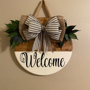 May include: A round wooden welcome sign with a white and brown finish. The sign has a burlap bow with black and white stripes and green leaves on either side. The sign says "Welcome" in black cursive lettering.