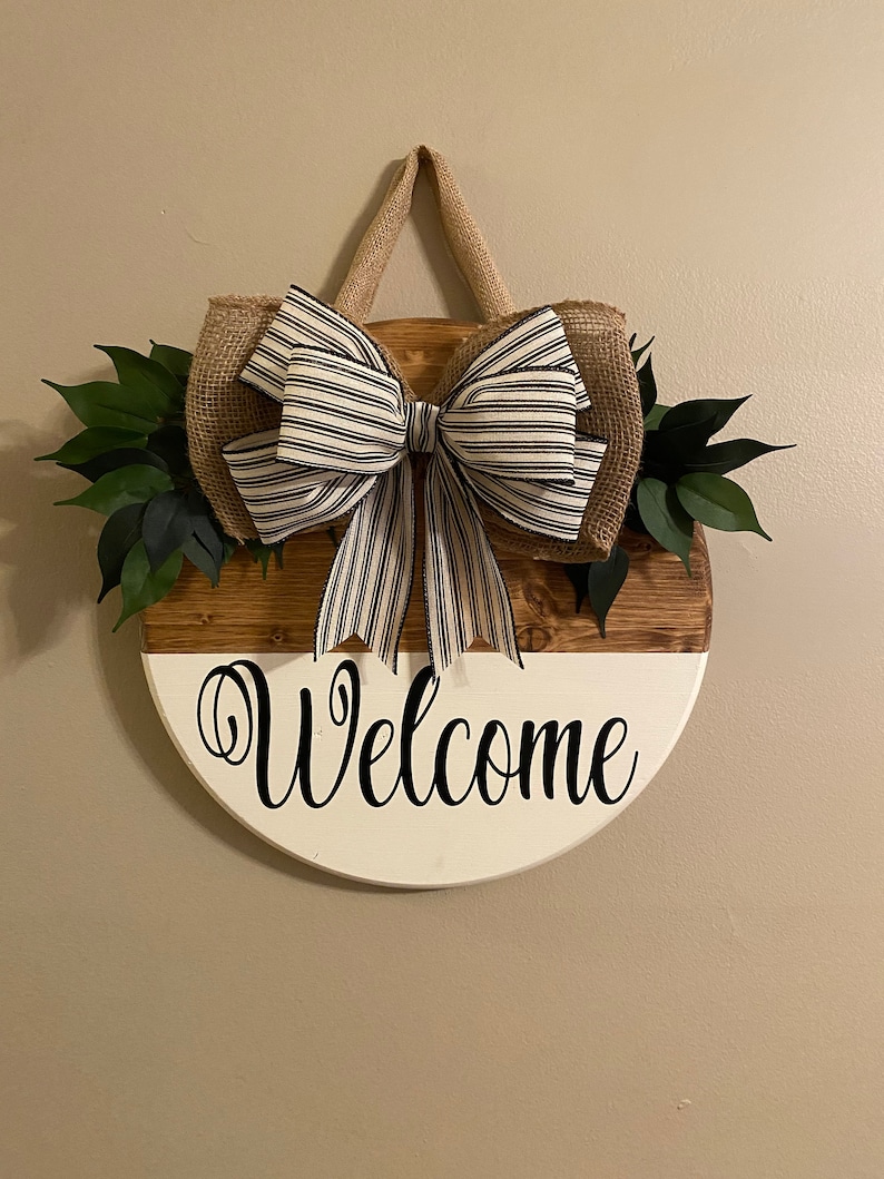 May include: A round wooden welcome sign with a white and brown finish. The sign has a burlap bow with black and white stripes and green leaves on either side. The sign says "Welcome" in black cursive lettering.