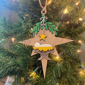 May include: A wooden Christmas ornament shaped like a star with a manger scene inside. The star is painted brown with a yellow star on top and green leaves around the edges. The manger is painted yellow with a white baby inside.